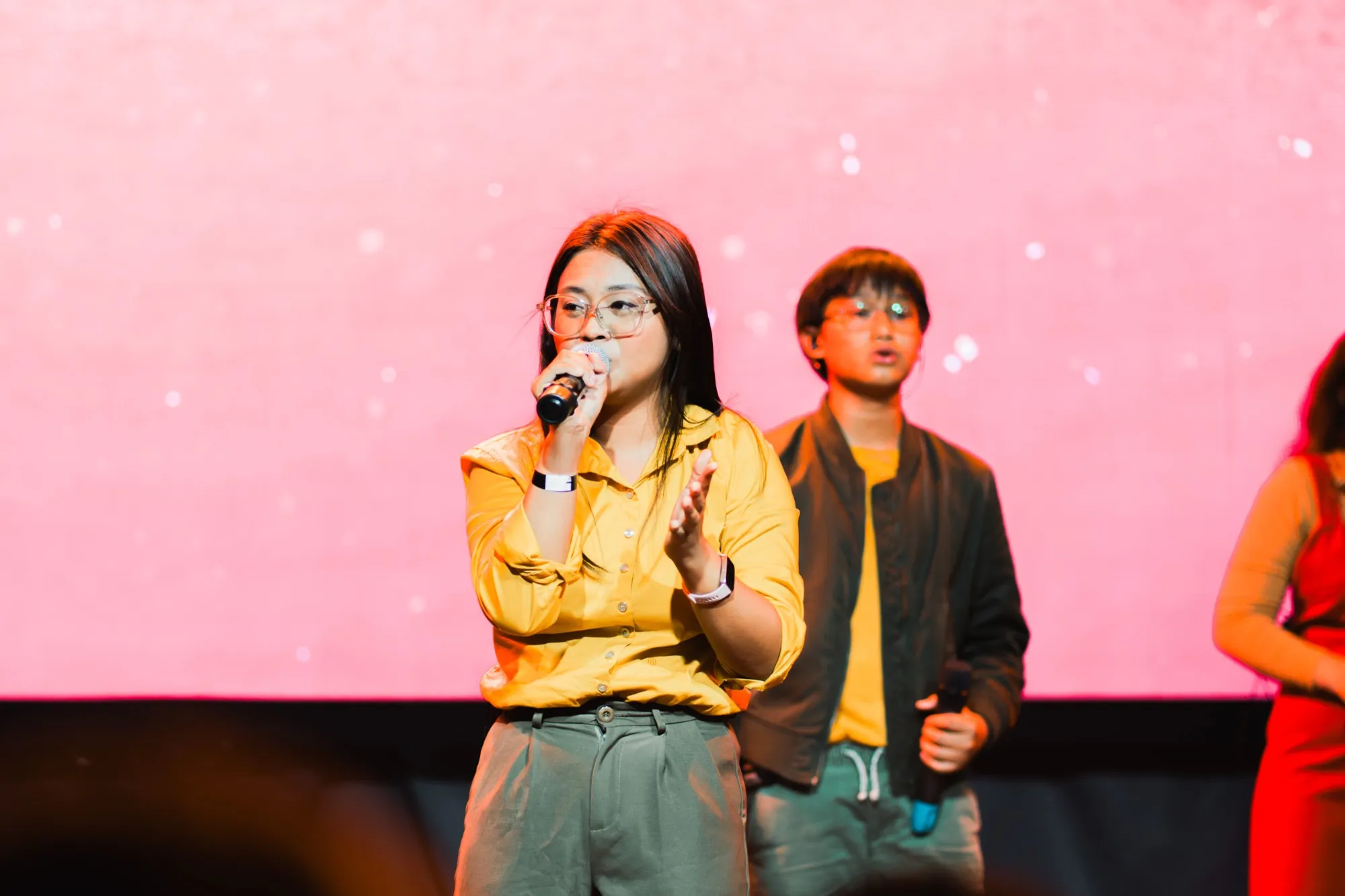 A singer in a yellow shirt and clear-framed glasses sings into a handheld microphone on stage, with two other vocalists blurred behind her against a pink LED wall