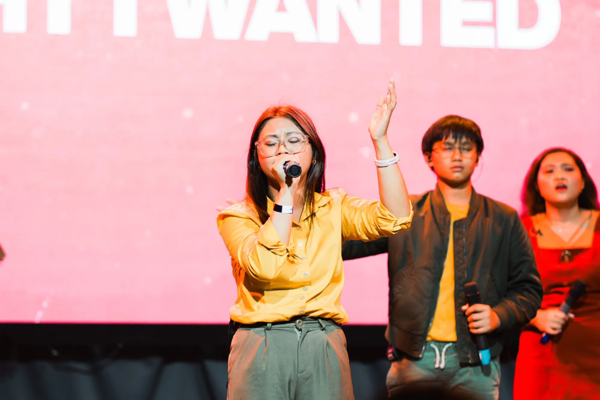 A vocalist sings with one hand raised while two backing singers stand behind her, in front of a pink screen with large white lettering