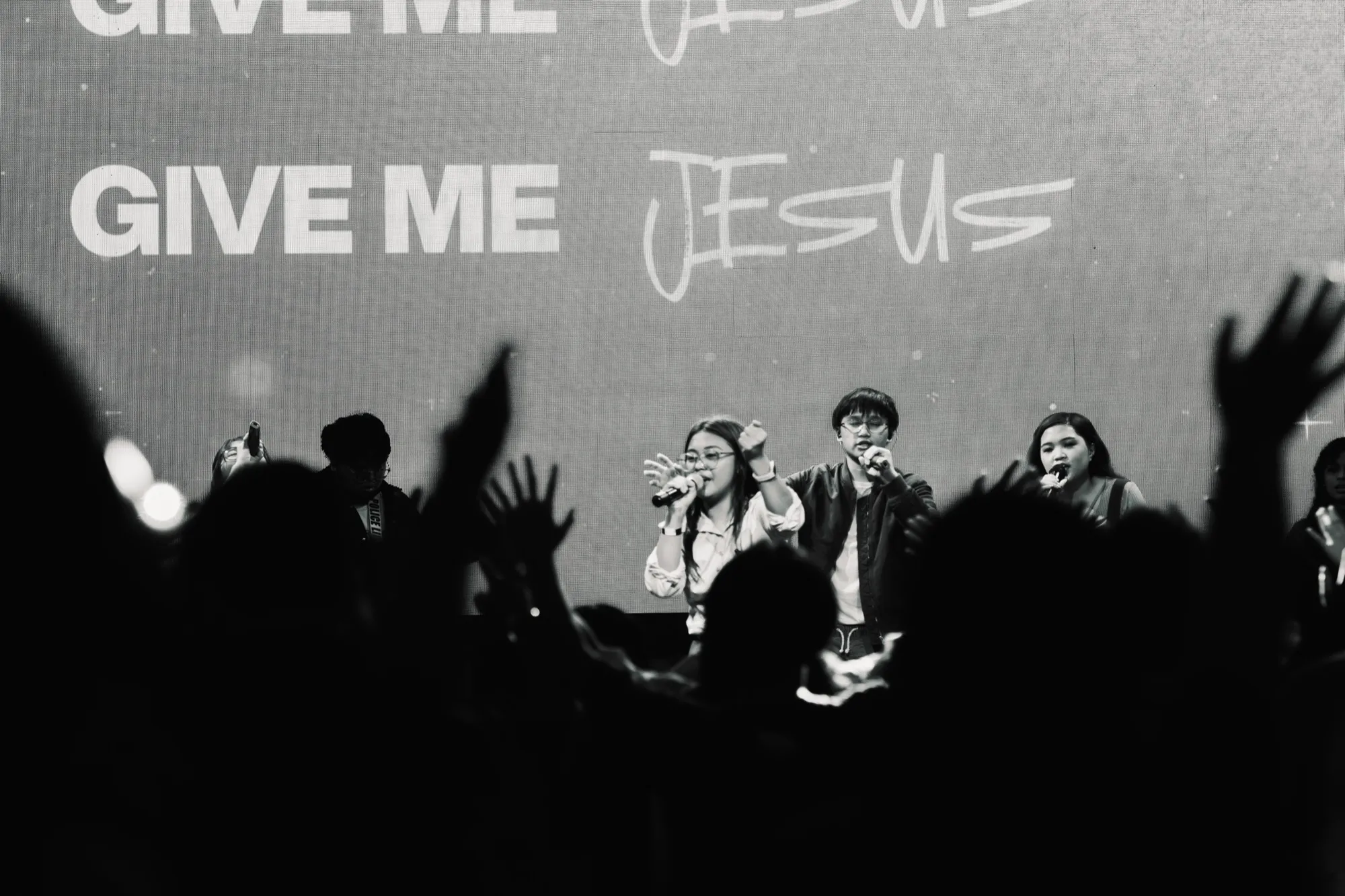 Black-and-white frame of three vocalists singing beneath a screen reading "GIVE ME JESUS", seen over the silhouetted raised hands of a congregation