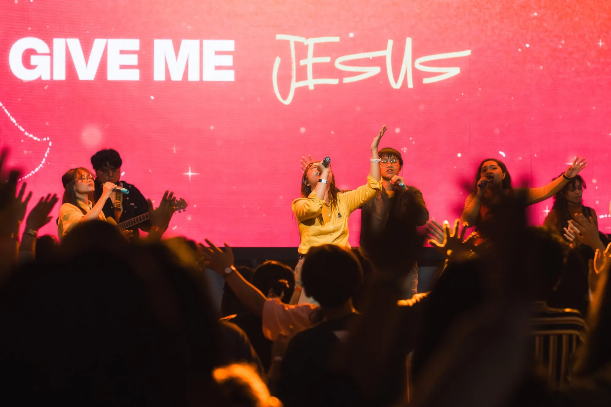 A worship band on a magenta-lit stage under a "GIVE ME JESUS" screen, photographed over a crowd of raised hands