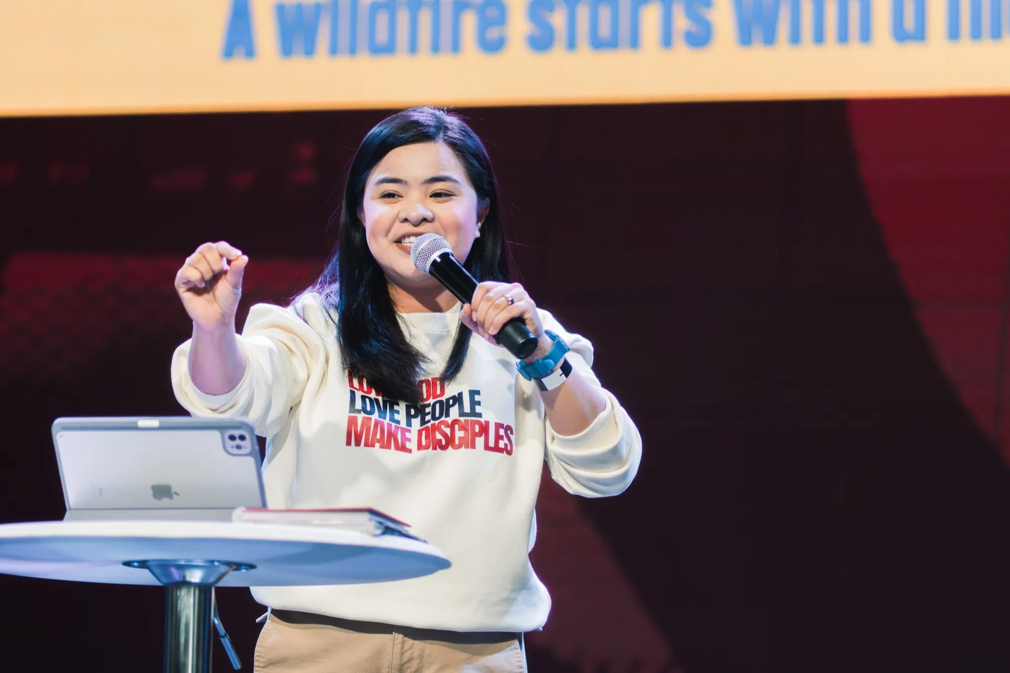 A speaker in a cream sweatshirt printed "LOVE GOD LOVE PEOPLE MAKE DISCIPLES" talks into a microphone beside a tablet on a small round table