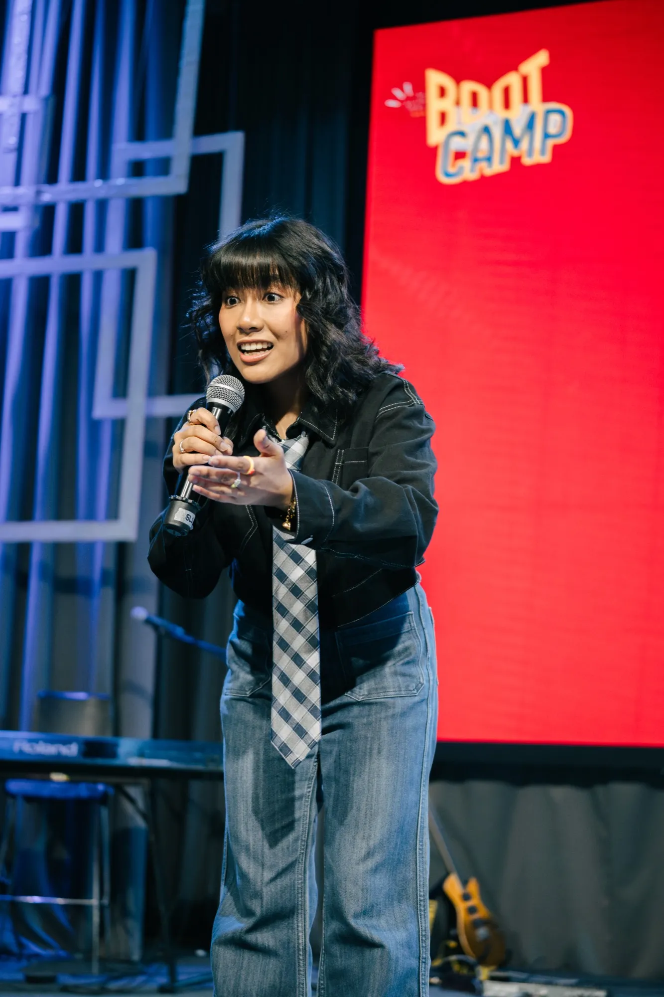 A host speaks into a microphone and gestures forward, with a red screen reading "BOOT CAMP" behind her