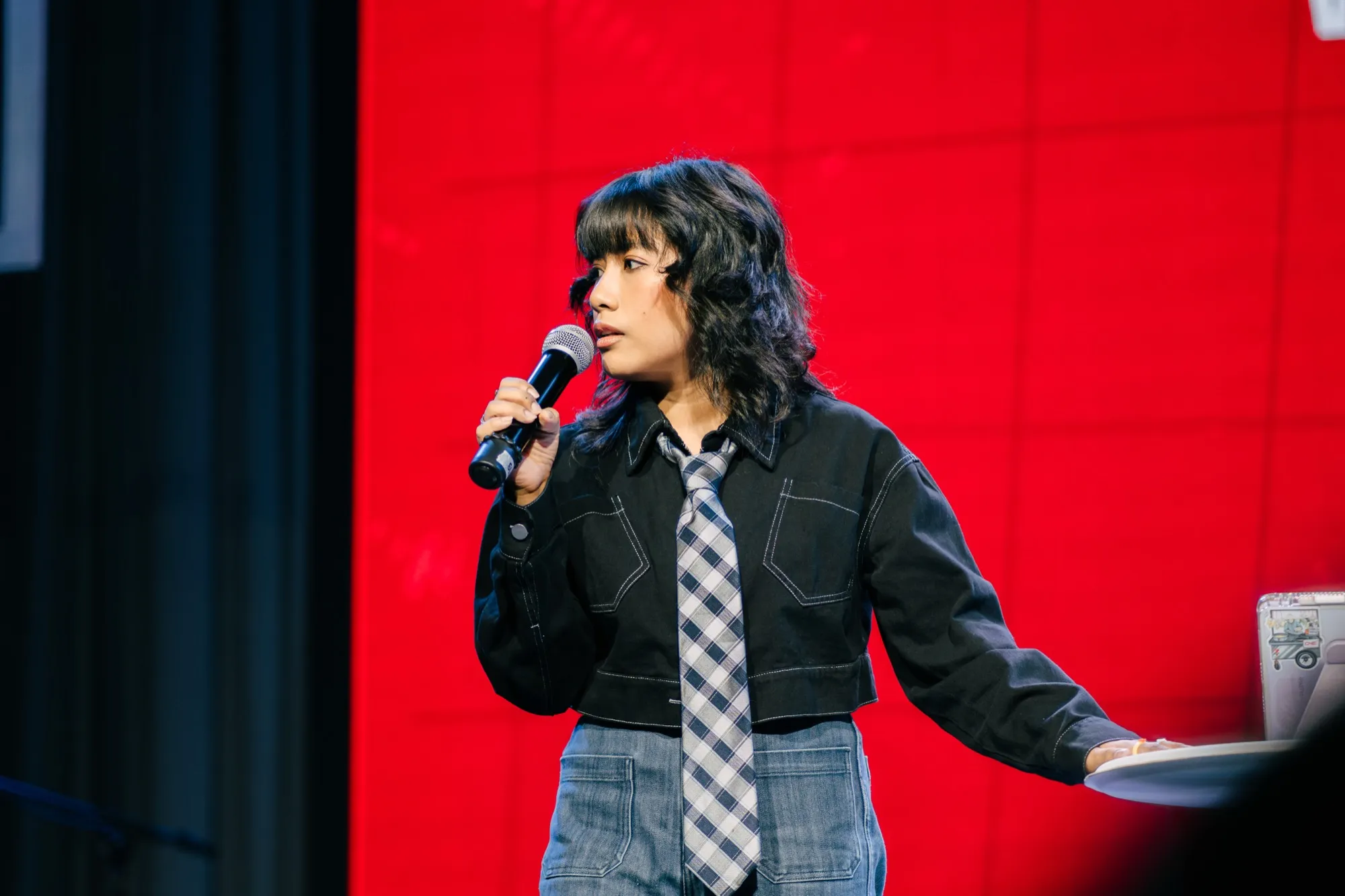 A host in a black jacket and checked tie speaks into a microphone, turned to her right, against a red screen