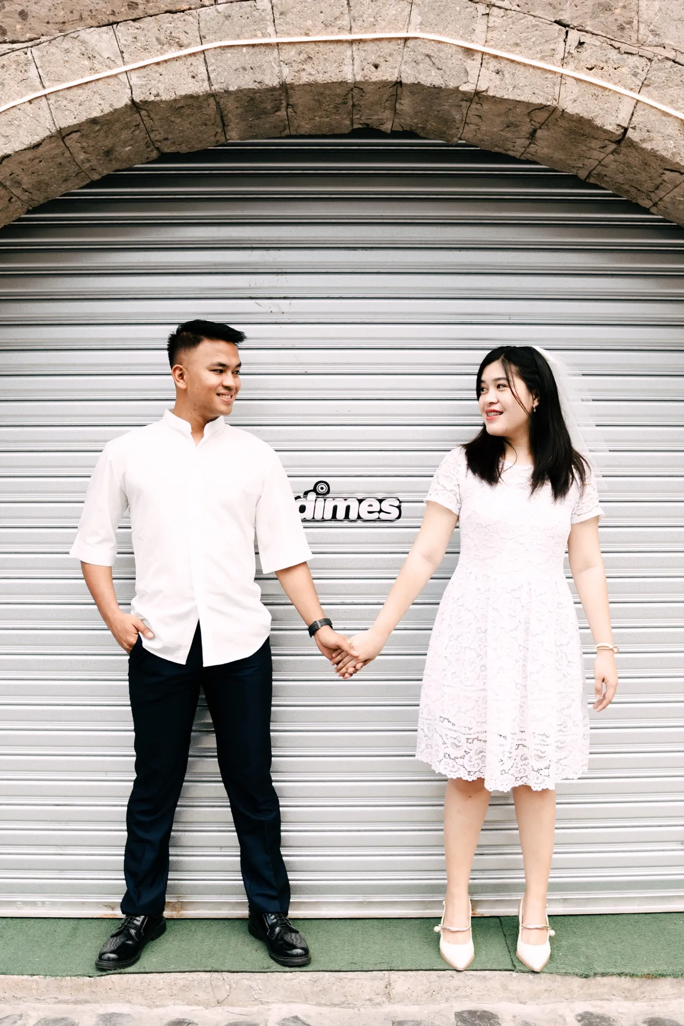 A couple in white stand apart holding hands in front of a corrugated metal roller shutter under a stone arch; she wears a white lace dress and short veil, he a white shirt and navy trousers