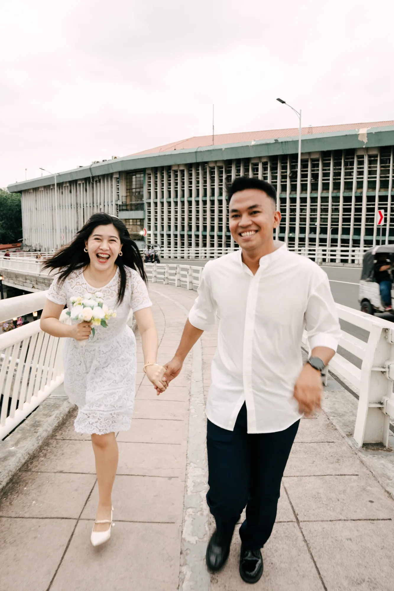 A couple walk hand in hand toward the camera on a bridge footpath, she holding a small white bouquet, with a louvred concrete building behind