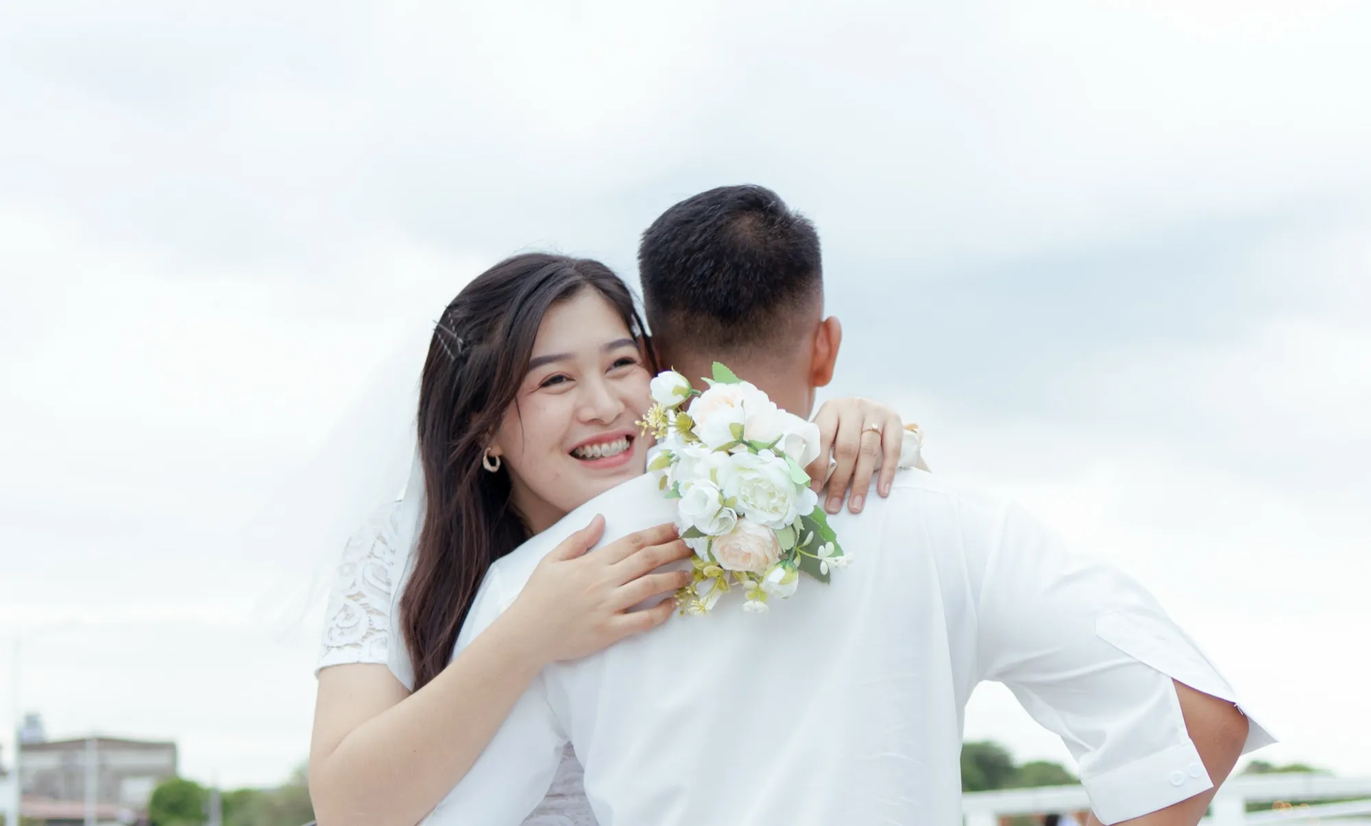 A woman in a white lace dress and veil rests her chin on a man's shoulder and looks at the camera, holding a white bouquet behind his back against an overcast sky