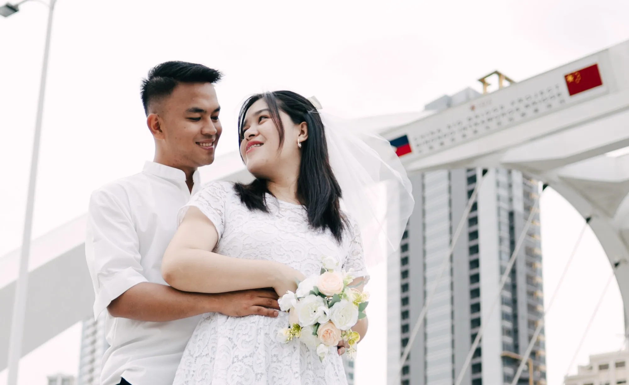 A man embraces a woman from behind on a bridge; she holds a cream-and-white bouquet, with the Binondo–Intramuros Bridge arch and high-rise towers behind