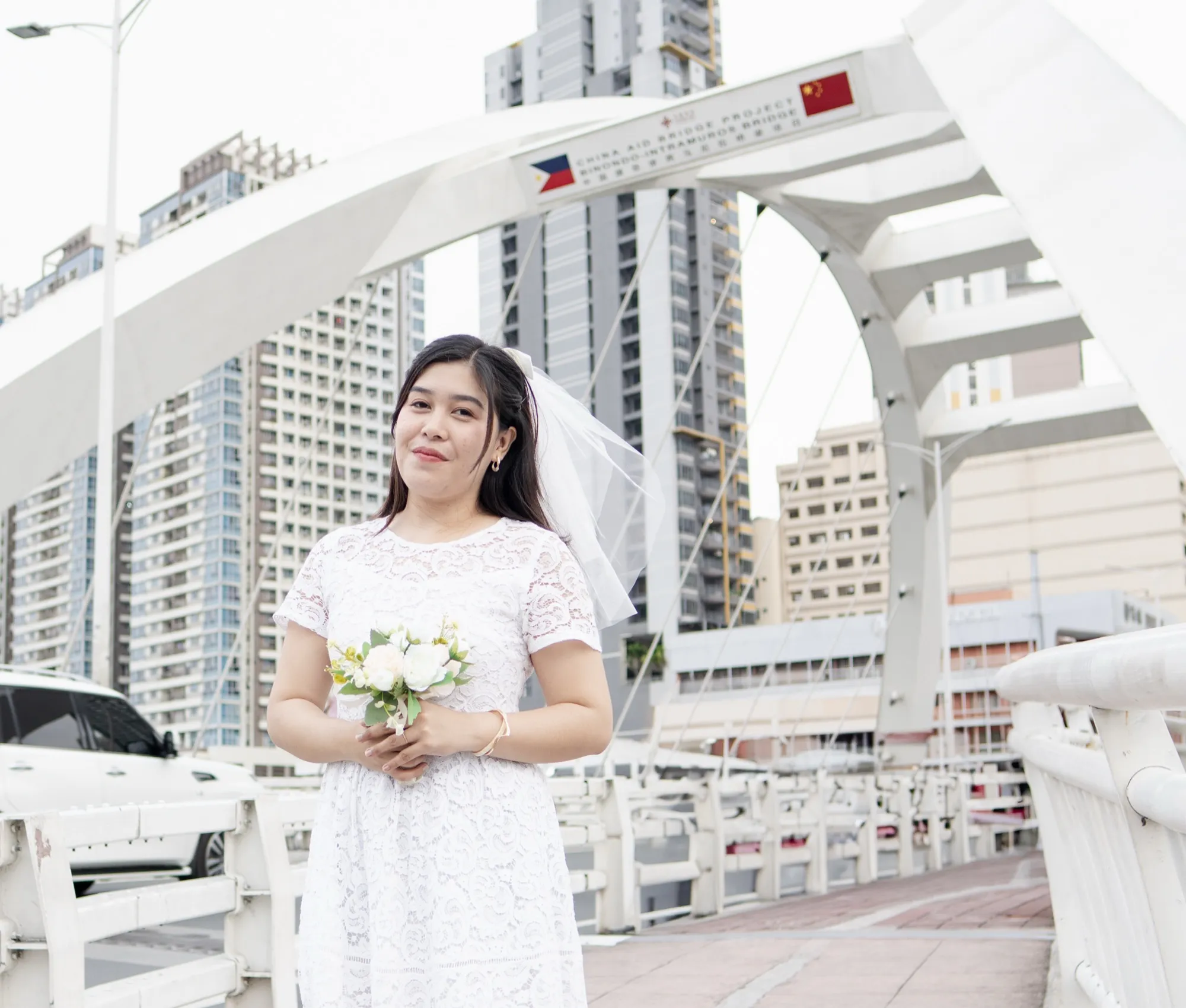 A woman in a white lace dress and veil holds a small bouquet while standing on the bridge, with the arch signed "Binondo–Intramuros Bridge" and city towers behind her