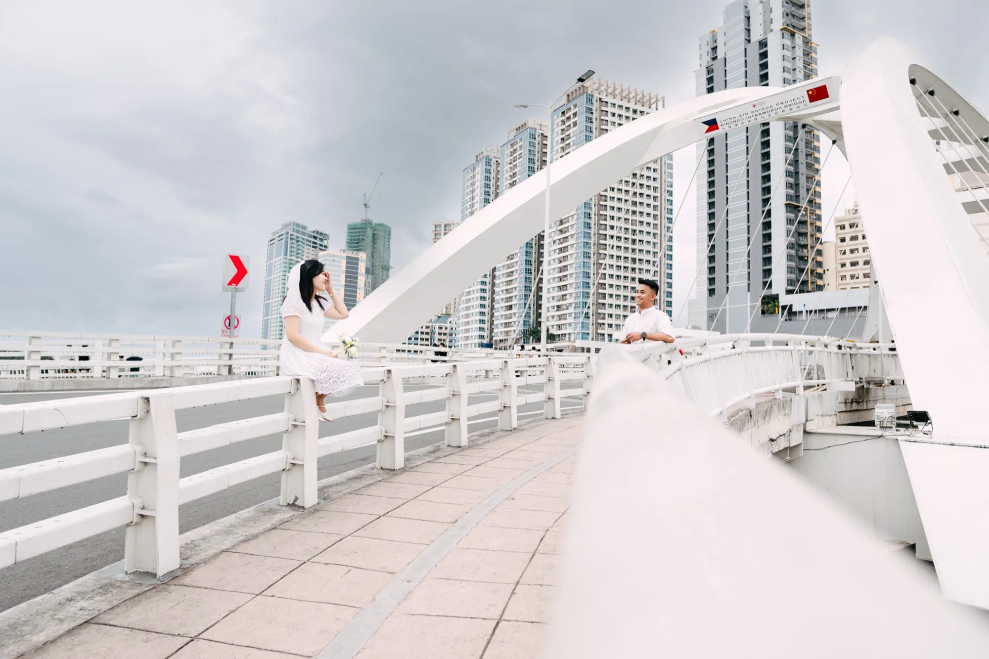 Wide view of a bridge walkway: a woman in white sits on the guardrail at left while a man in a white shirt leans on the railing at right, with the white bridge arch and city skyline behind