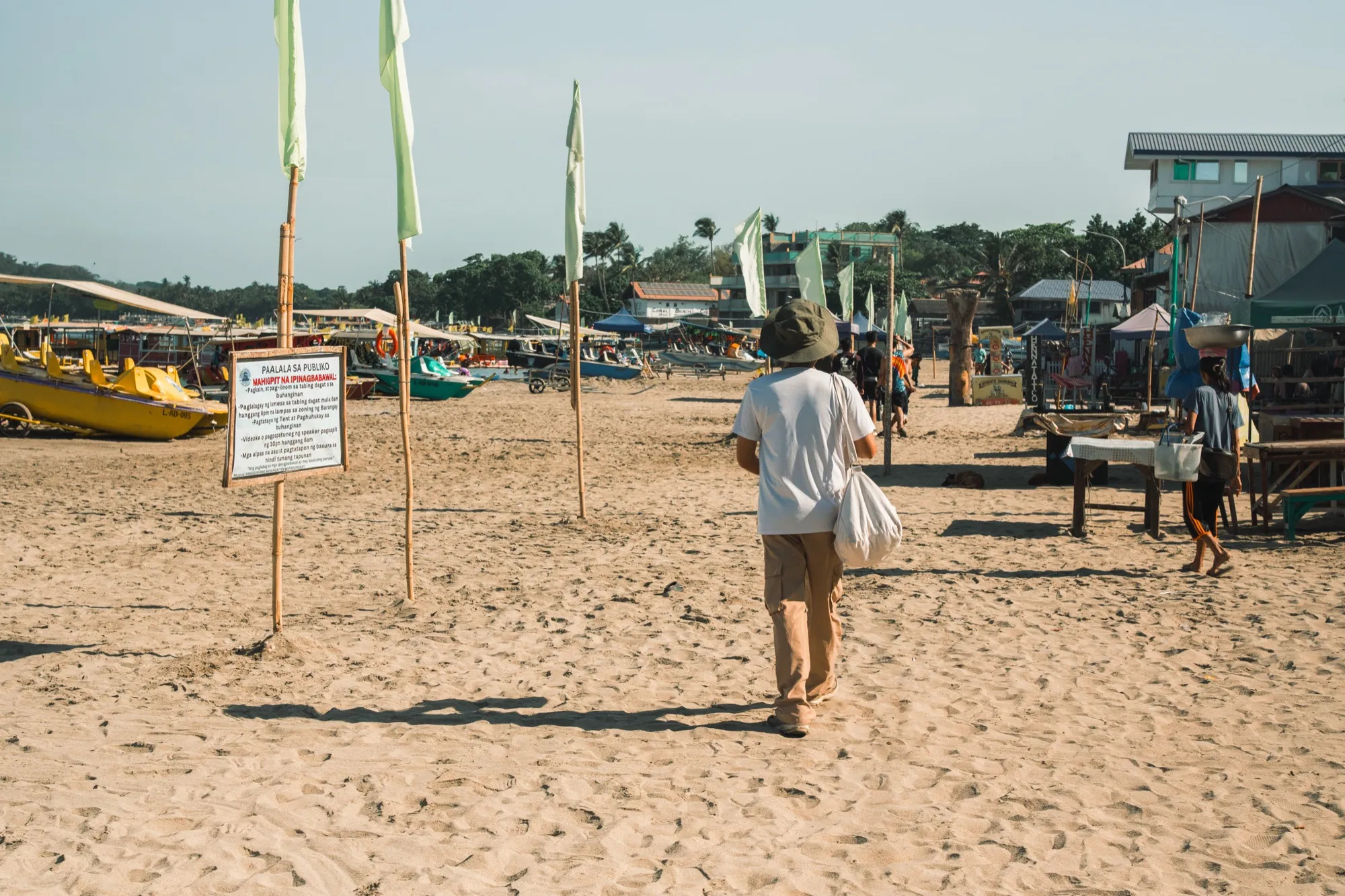A person in a bucket hat carrying a duffel walks away across a busy beach past bamboo flag poles, moored boats and beachfront stalls