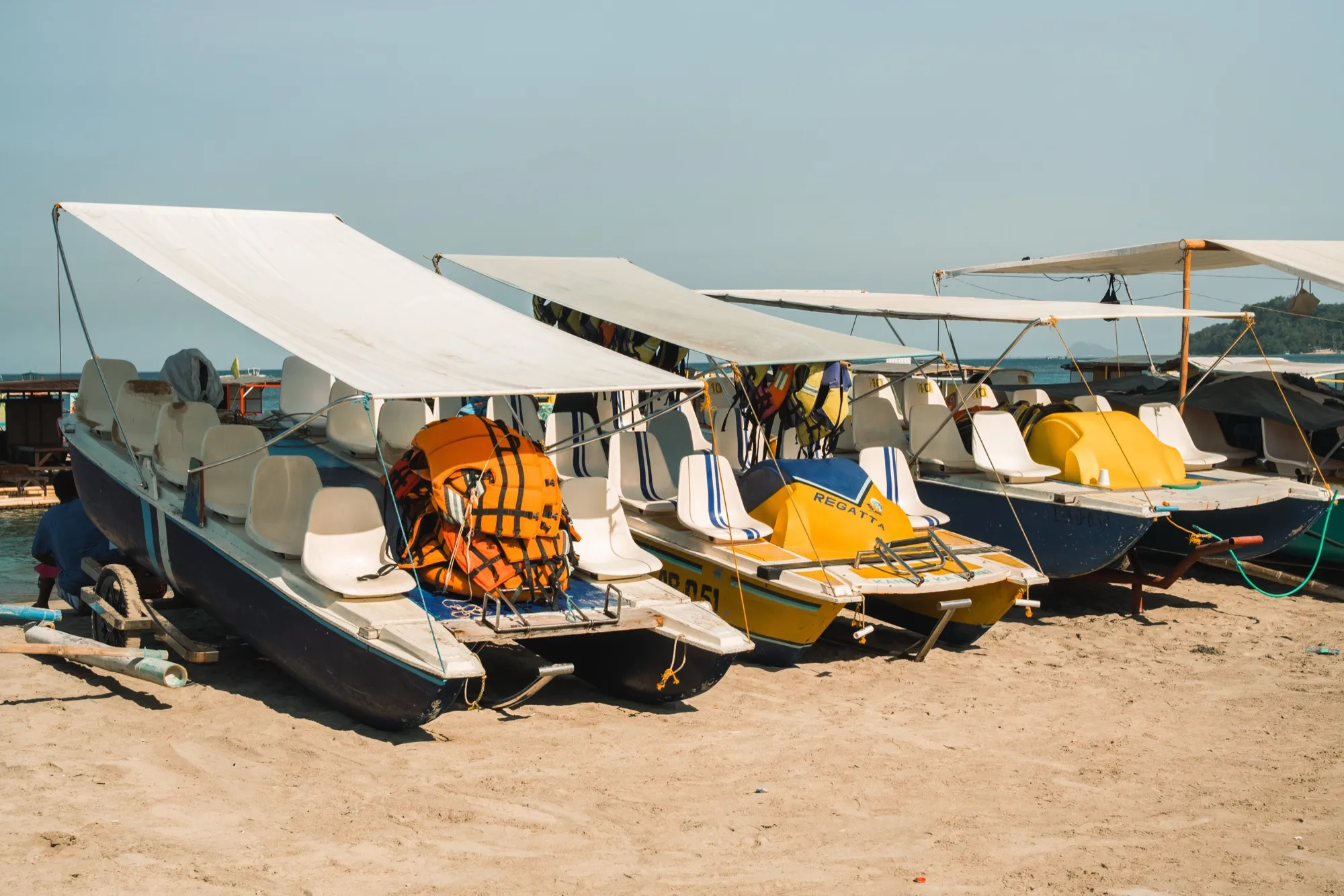 A row of canopied pedal boats with white seats and life vests parked on pale sand under a hazy sky