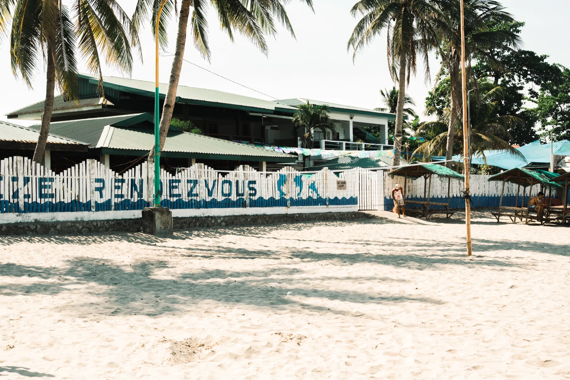 A white and blue painted fence reading "RENDEZVOUS" fronts a green-roofed beachfront building framed by palm trees, with empty white sand in the foreground