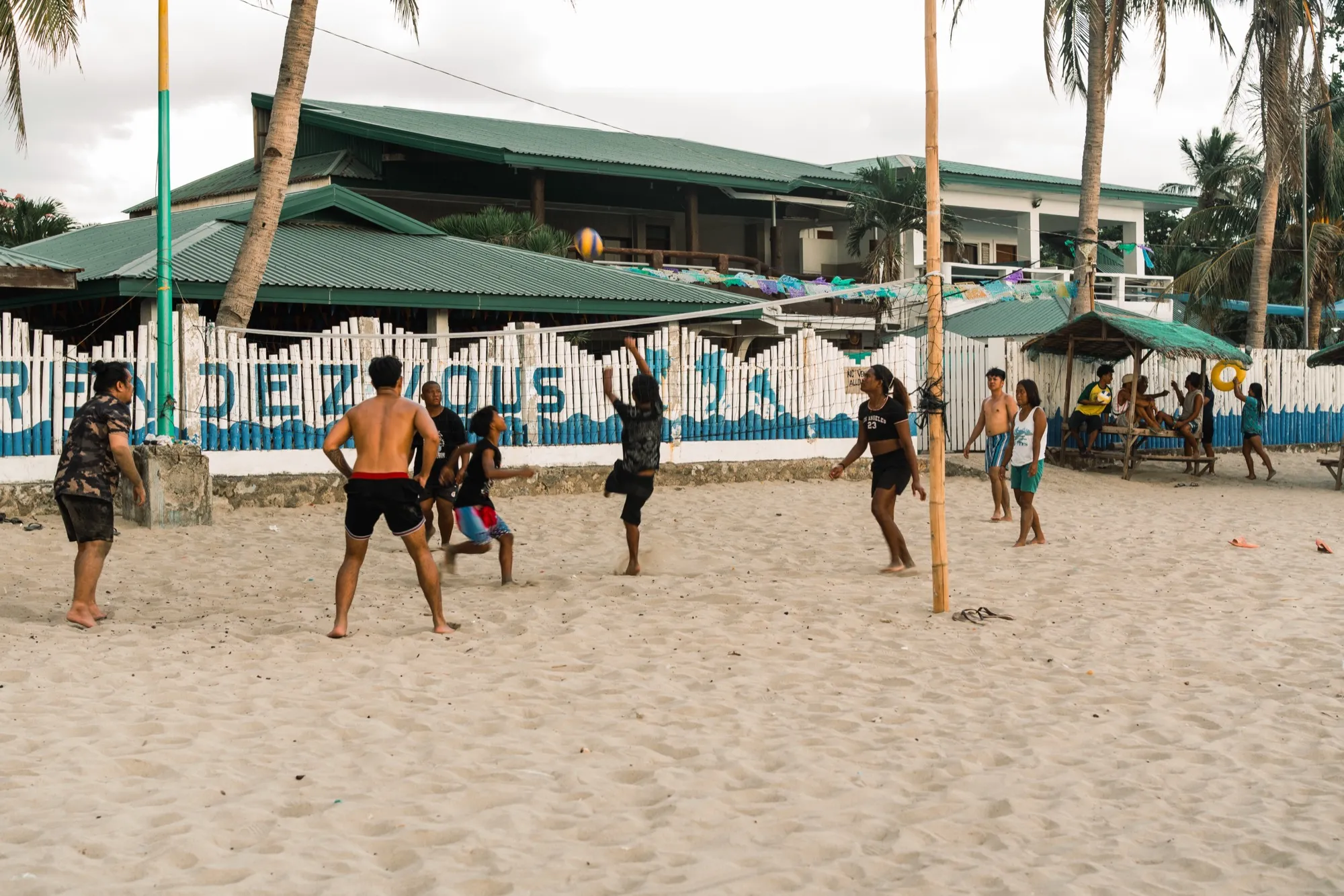 People play beach volleyball on white sand in front of the painted "RENDEZVOUS" fence, with onlookers seated at a thatched hut