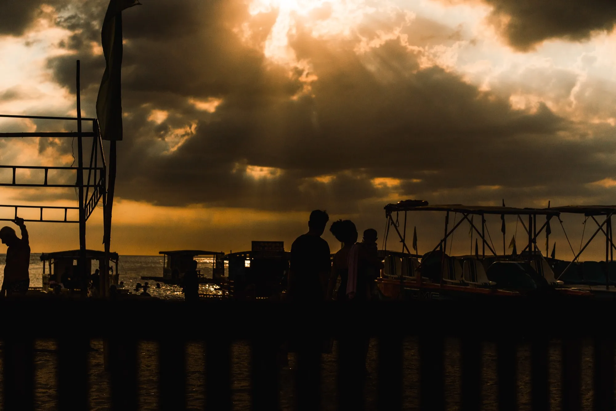 Silhouetted figures and moored boats against a cloudy sunset over the sea