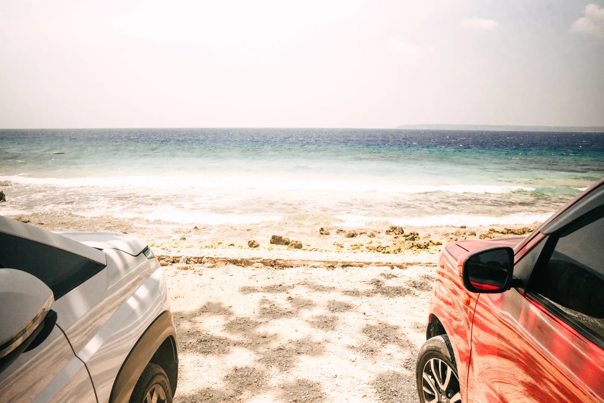Two parked cars frame a view of a rocky shoreline and clear turquoise sea under a bright sky
