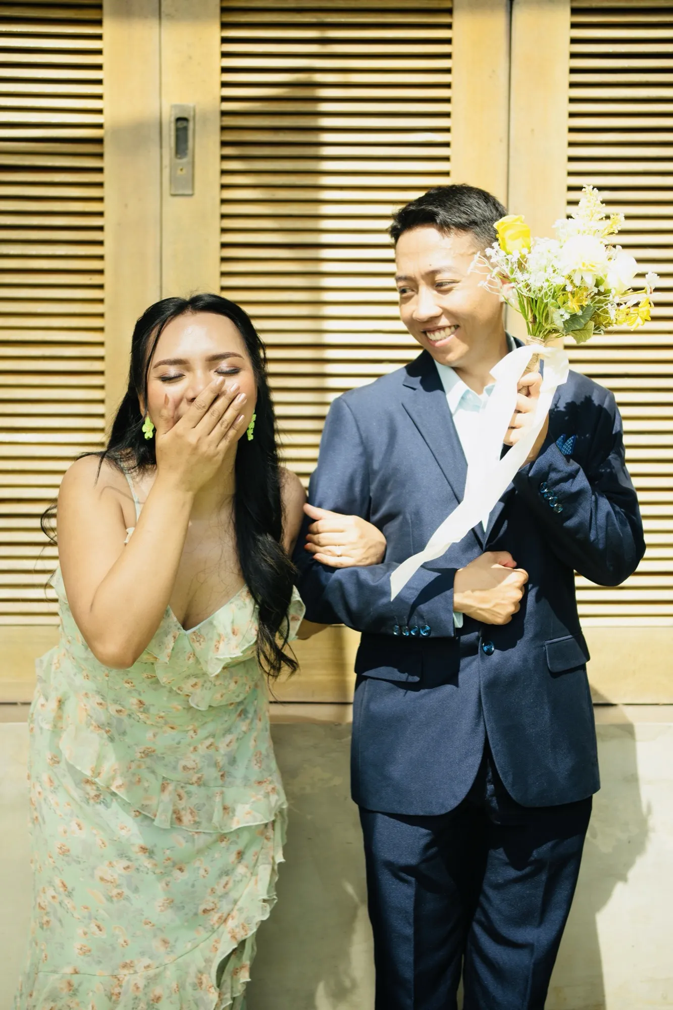 A woman in a pale green floral dress covers her mouth with her hand while a man in a navy suit holds a yellow-and-white bouquet beside her, in front of louvred wooden doors
