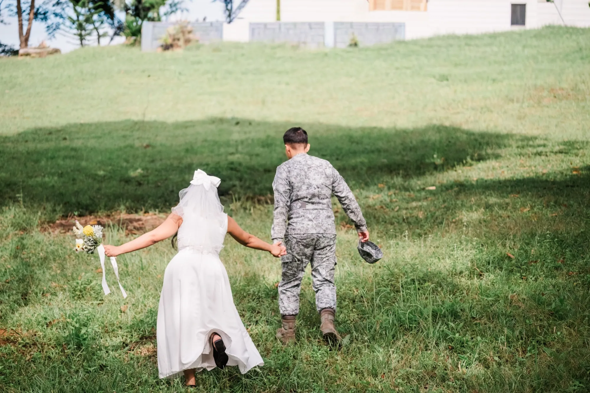 A woman in a white gown and veil and a man in camouflage uniform walk hand in hand up a grass slope away from the camera, he carrying his cap