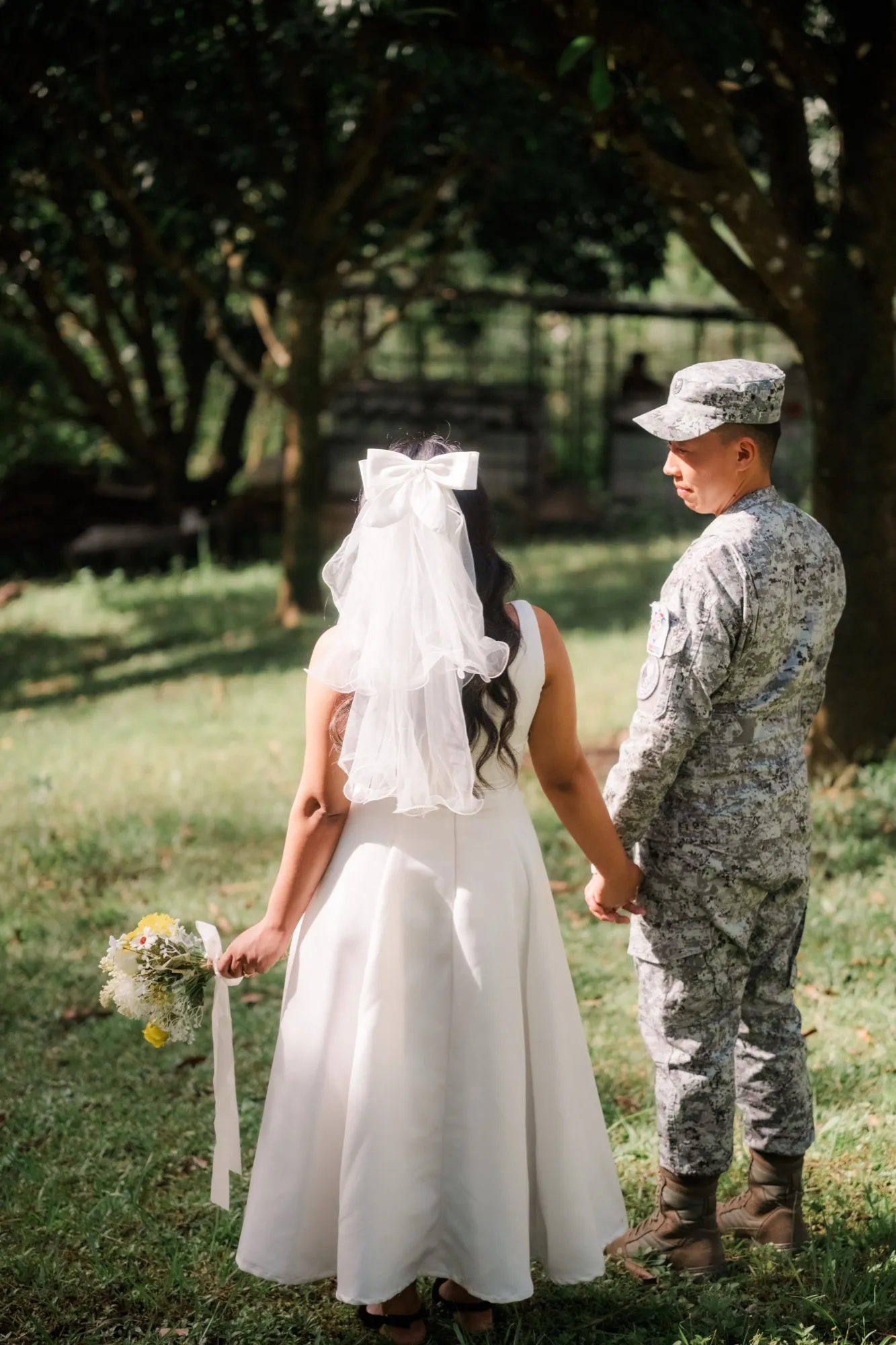 A woman in a white gown and bow-tied veil stands with her back to the camera holding hands with a man in camouflage uniform and cap, under trees