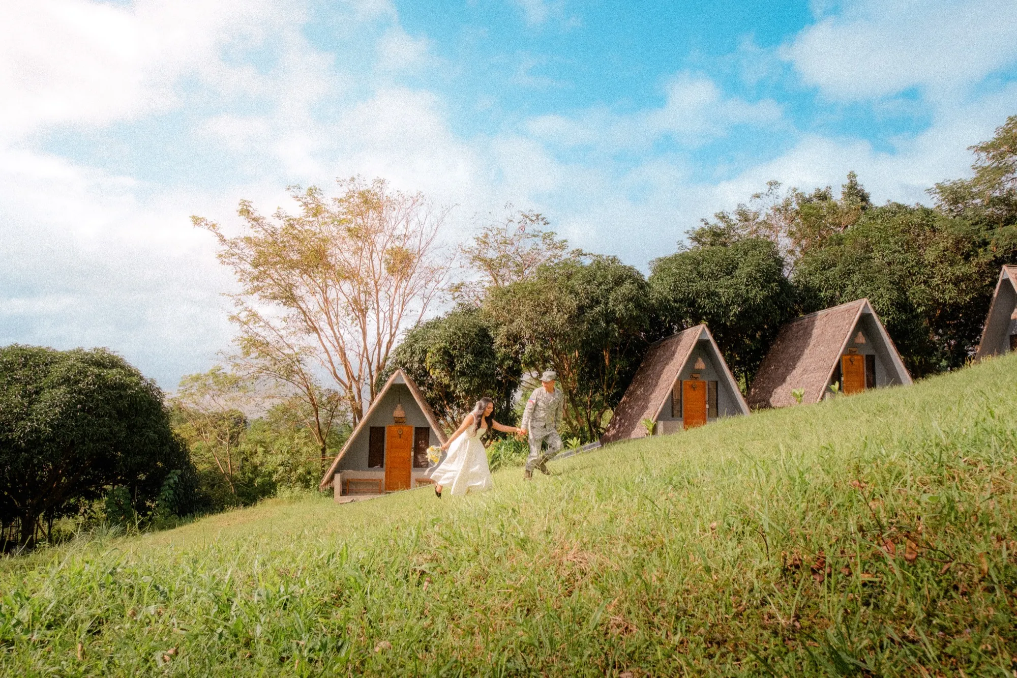 A couple walk hand in hand up a grassy hillside past a row of A-frame cabins under a blue sky with clouds