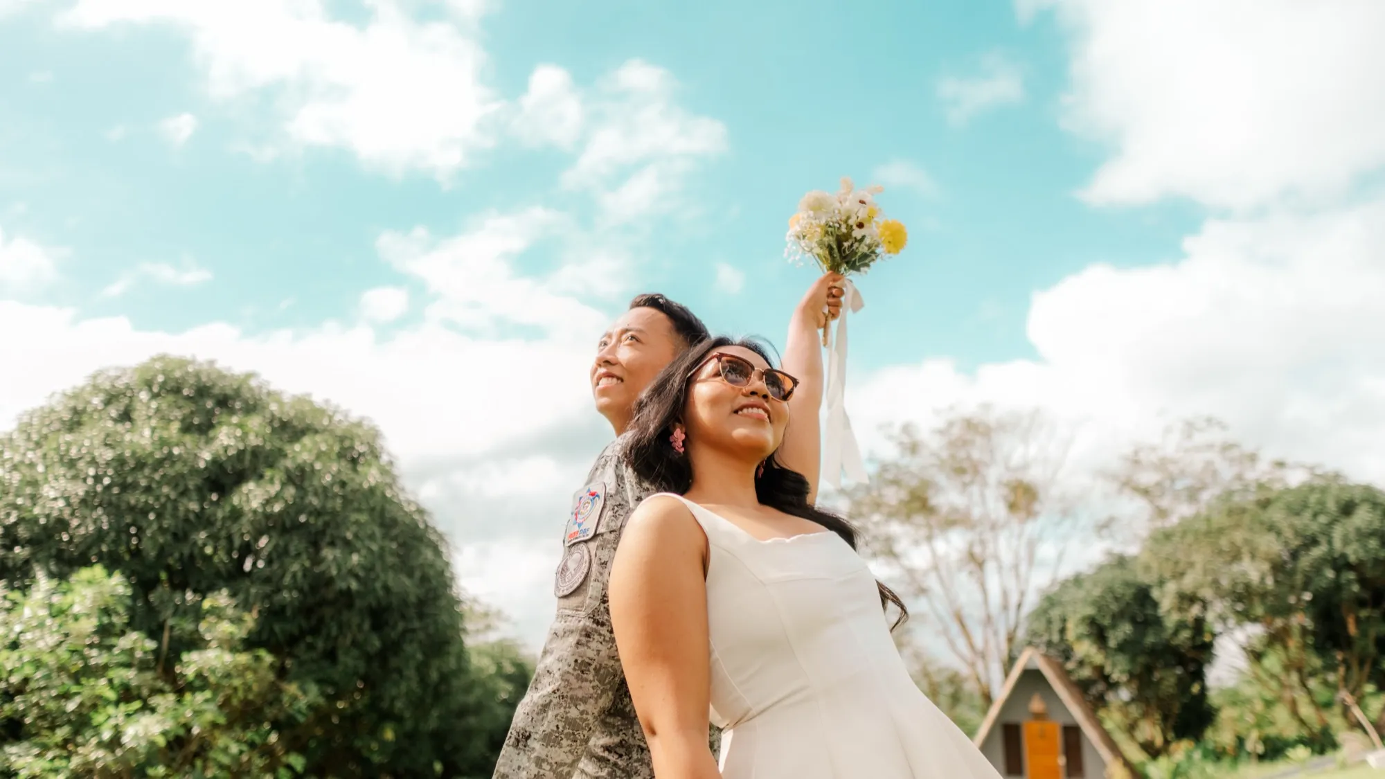 A woman in a white dress and sunglasses raises a bouquet overhead while standing back to back with a man in camouflage uniform against a bright cloudy sky