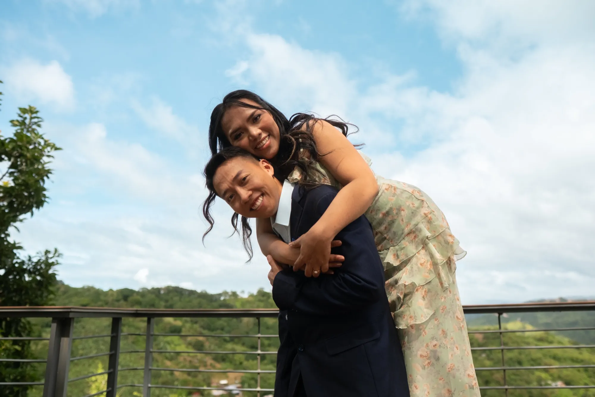 A woman in a green floral dress rides piggyback on a man in a navy suit on a railed deck, both looking at the camera, with green hills behind