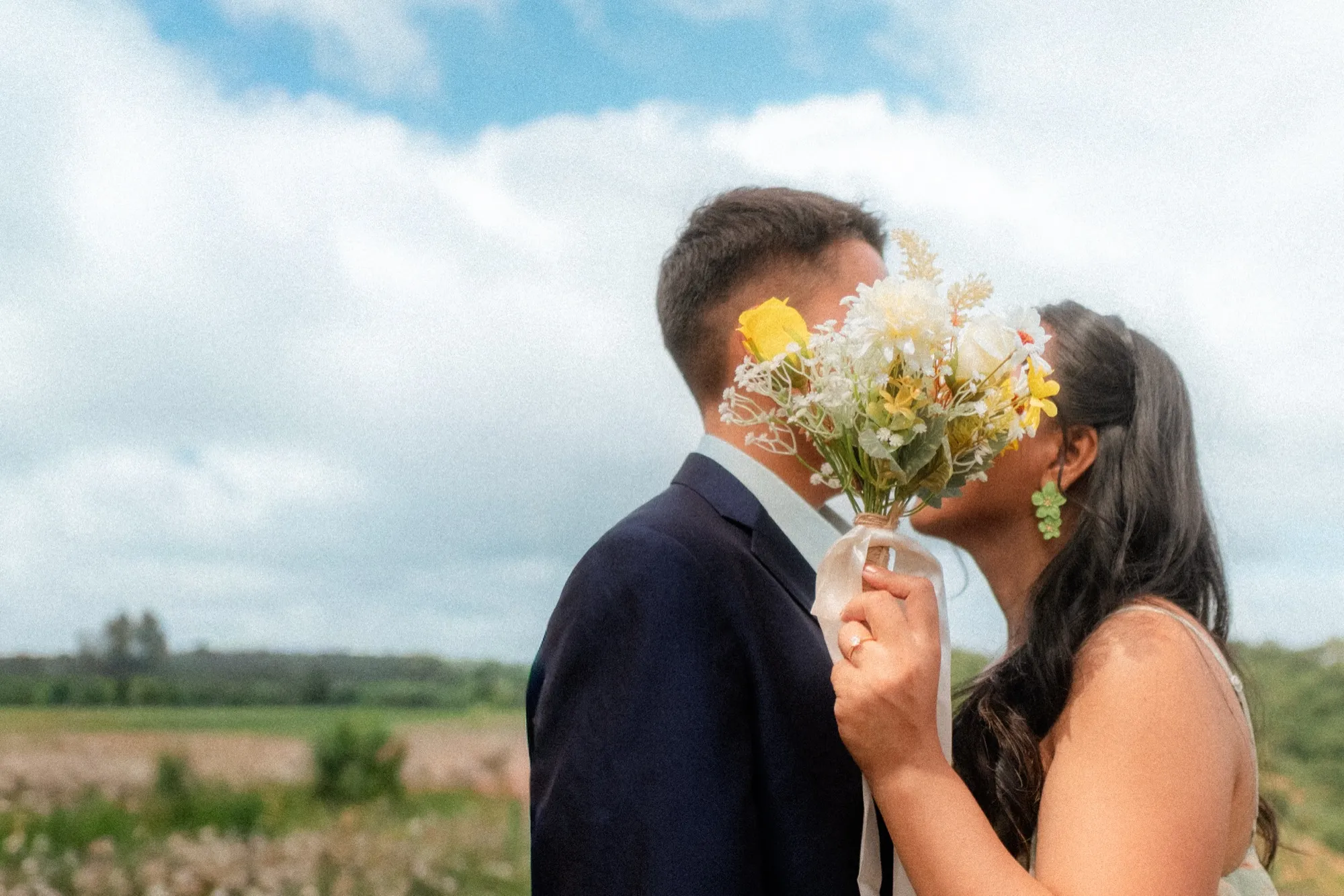 A couple kiss behind a raised yellow-and-white bouquet that hides their faces, standing in an open field under a cloudy sky