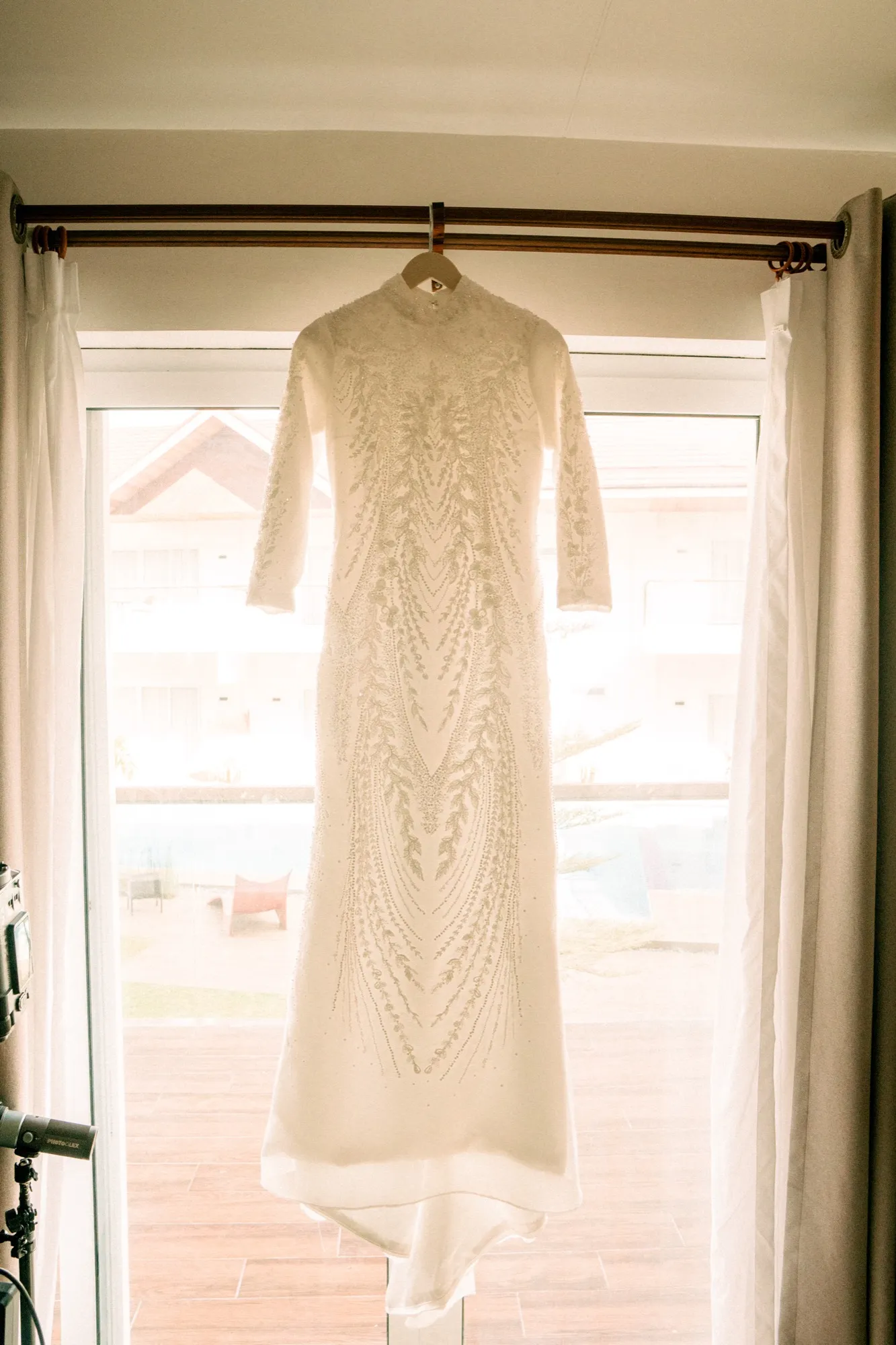 A beaded long-sleeved white wedding gown hangs from a curtain rod in front of a bright window overlooking a poolside courtyard