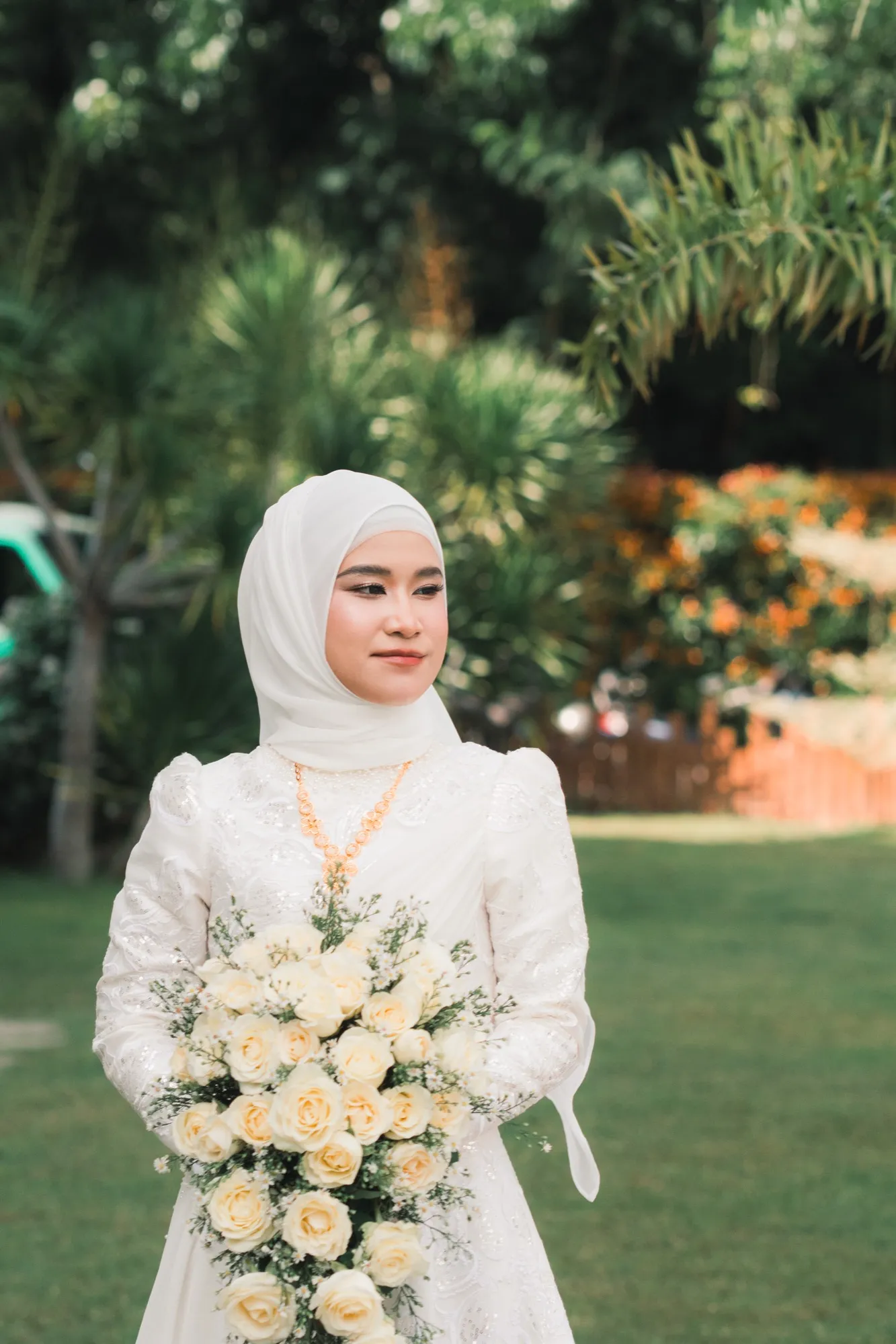 A bride holds a cascading bouquet of cream roses on a lawn with palms and orange-flowering shrubs behind her