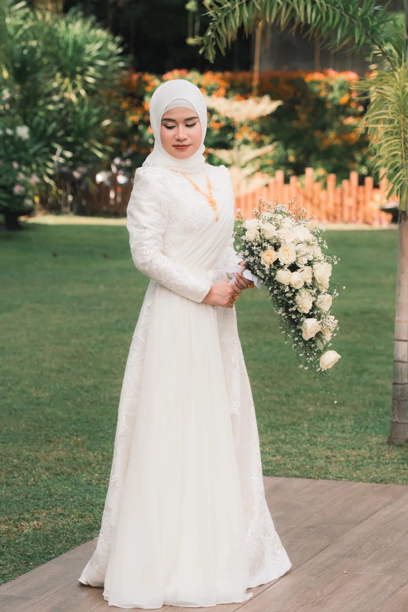 A bride stands on a wooden walkway on a lawn, looking down at the cascading bouquet held at her side