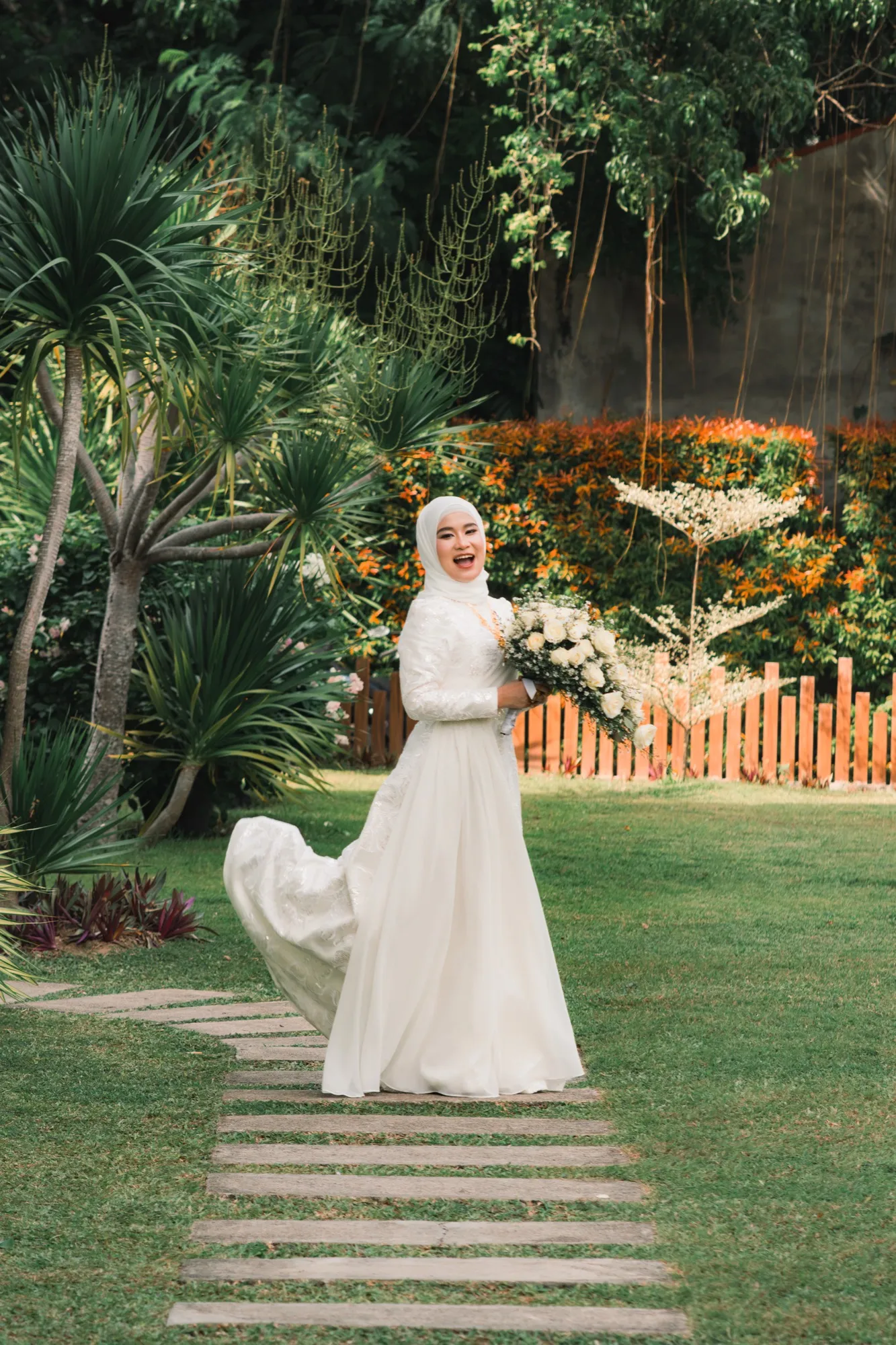 A bride laughs on a stepping-stone path in a garden, her gown train sweeping behind her, bouquet held out to one side