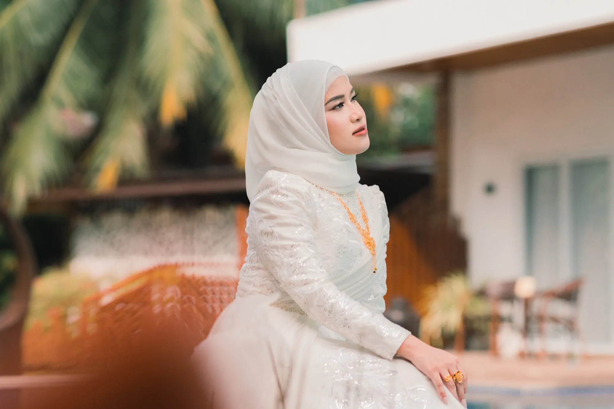 A bride sits in profile beside a pool, wearing a white hijab, beaded gown and gold necklace, with palms and a garden wall blurred behind