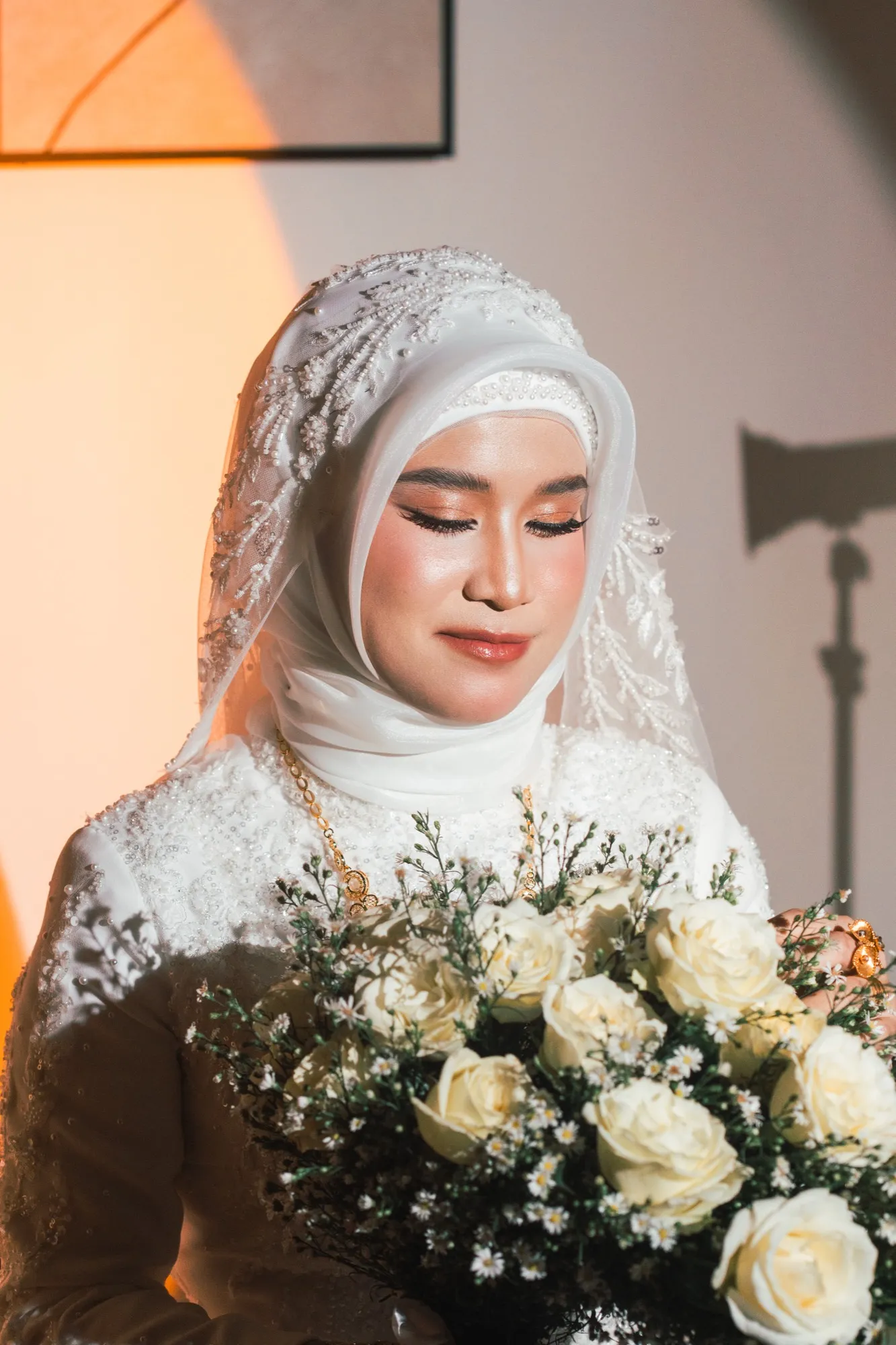 A bride in a beaded veil and hijab looks down at a large bouquet of cream roses, lit against a warm interior wall