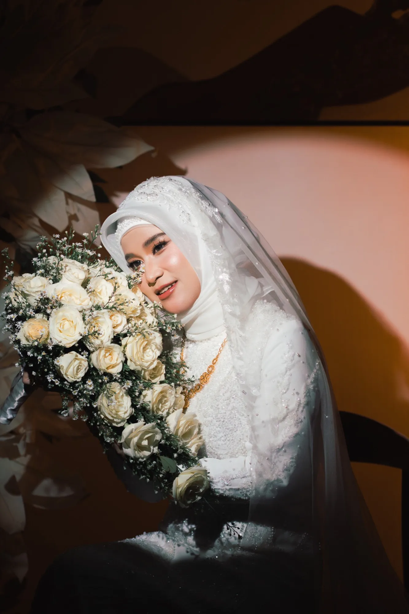 A bride leans toward her bouquet of cream roses, veil draped over her shoulder, against a warm-lit wall