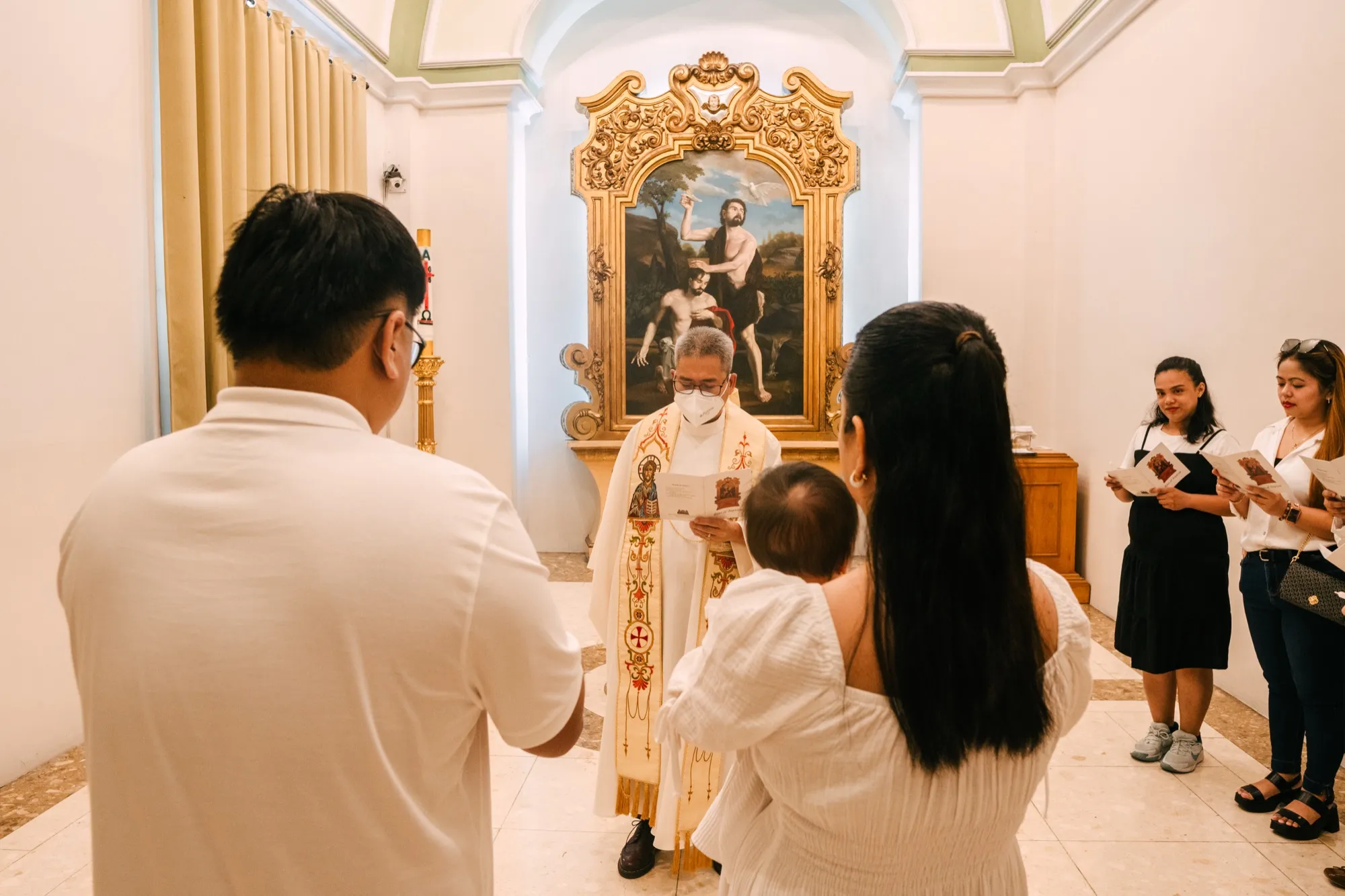 A priest in white vestments reads from a booklet before a gilded altarpiece while a couple seen from behind holds an infant, with guests reading along at right