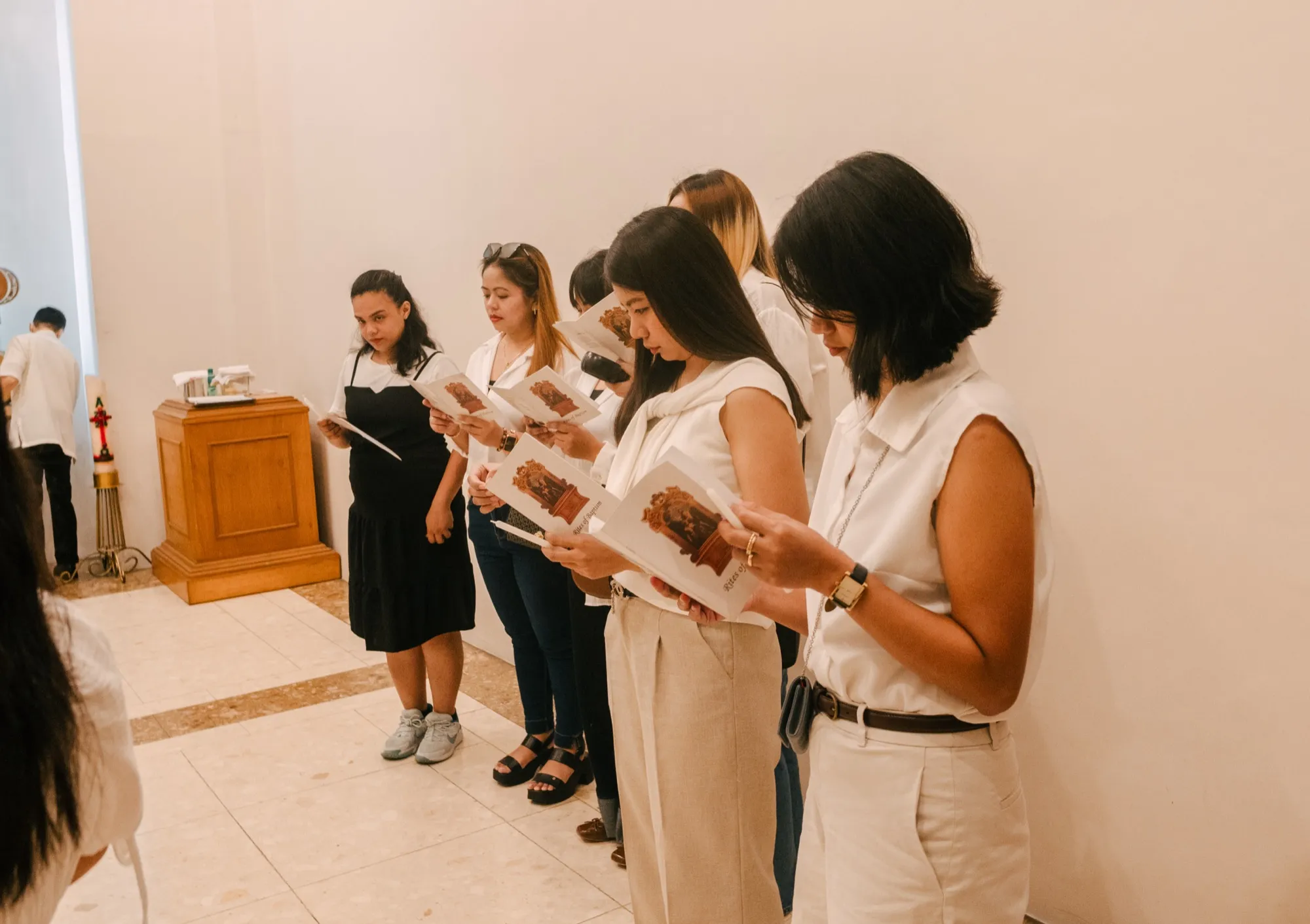 A row of guests in white and cream stand along a chapel wall reading from Rites of Baptism booklets