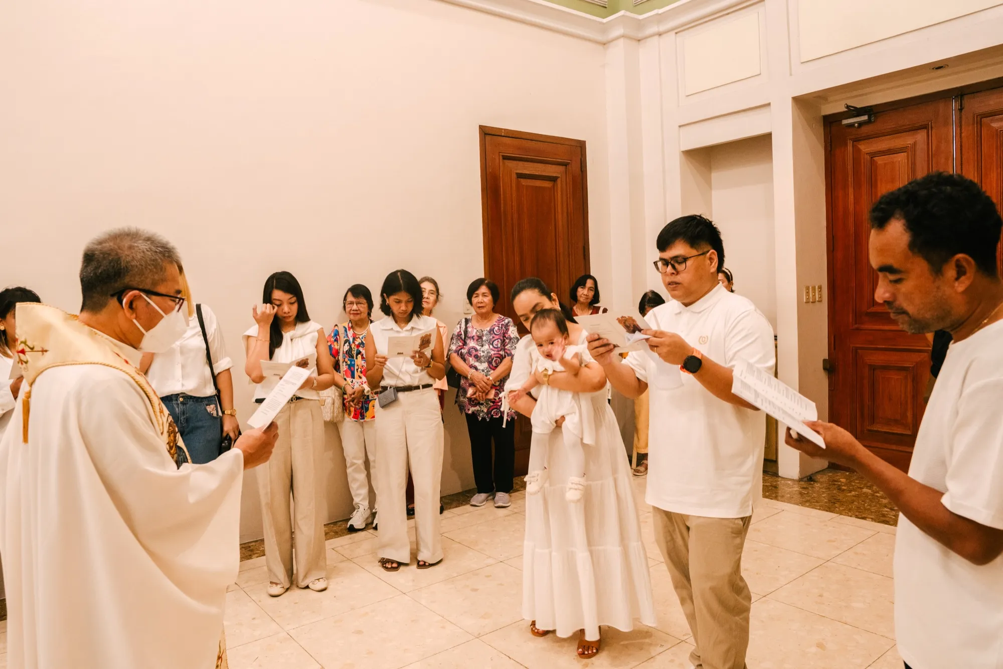 A priest reads from a booklet at left as a woman holding an infant, a man in a white polo and a group of guests stand in a chapel vestibule