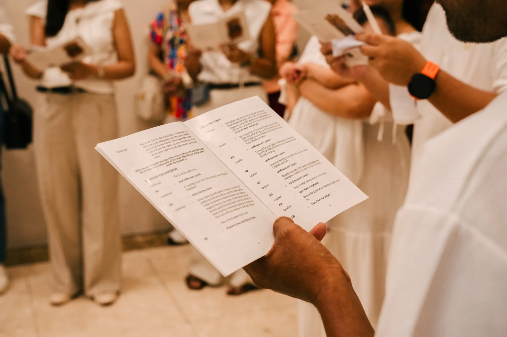 A hand holds an open Rites of Baptism booklet showing the Gospel and intercessions, with blurred guests behind