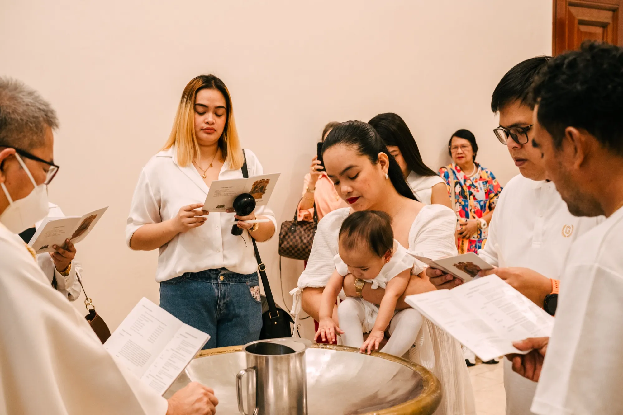A woman lowers an infant toward a gilded baptismal font as a priest and guests read from booklets around them