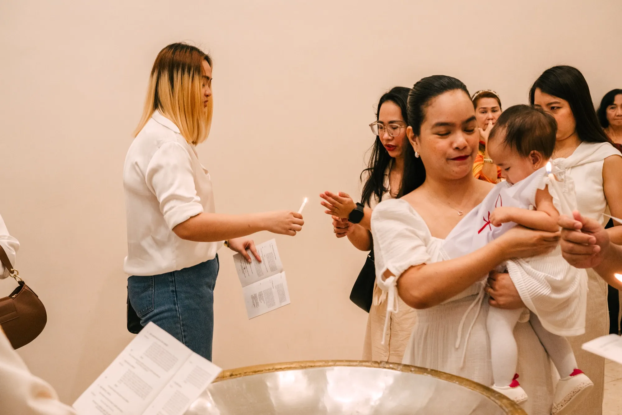 A woman in a white shirt passes a lit taper across the baptismal font toward a woman holding an infant