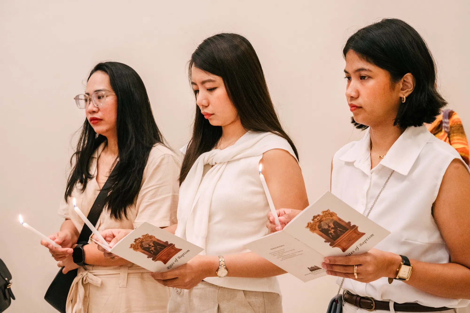 Three guests in white and cream stand holding lit candles and open Rites of Baptism booklets